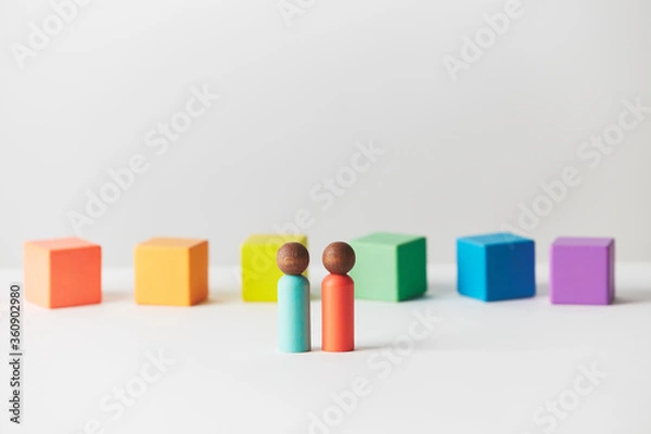 Fototapeta Wooden figures concept. Couple standing in front of wooden blocks in colours of the gay pride rainbow flag. Couple of dark skin colour. 