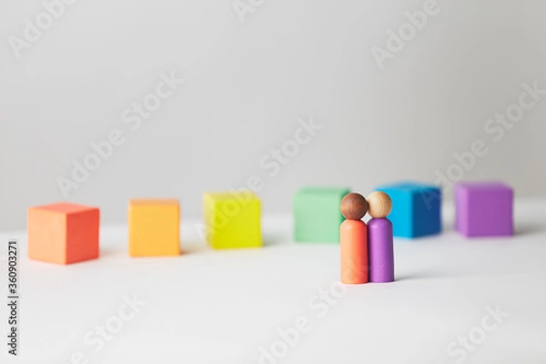 Fototapeta Wooden figures concept. Couple standing in front of wooden blocks in colours of the gay pride rainbow flag. Couple of mixed skin colour. 