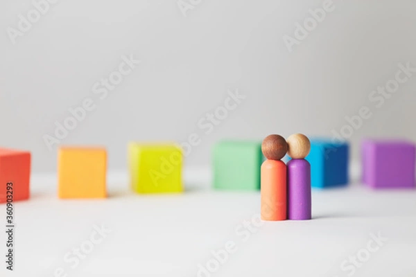 Fototapeta Wooden figures concept. Couple standing in front of wooden blocks in colours of the gay pride rainbow flag. Couple of mixed skin colour.
