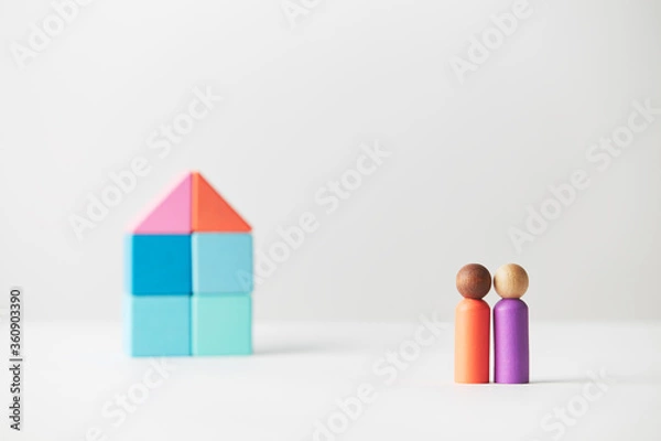 Fototapeta Wooden figurines concept. Two wooden figures people standing in front of a large multi generations block house, mixed race skin colour, four adults. Realty market.