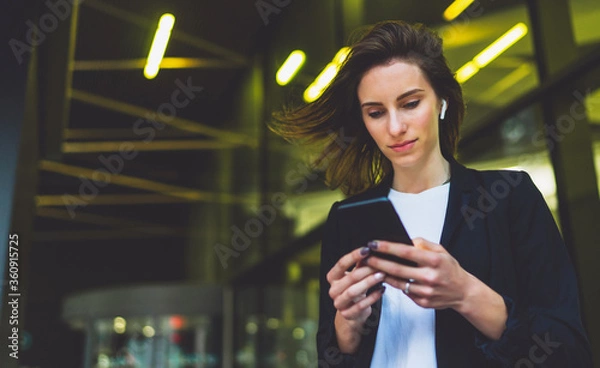 Fototapeta concentrated businessman wearing black suit using modern smartphone near office, successful employer chats mobile phone while standing near skyscraper office on background nigh flare light