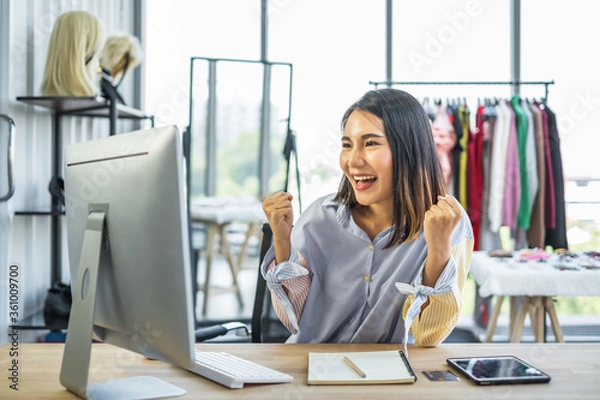 Obraz Young Asian woman business owner sitting at a desk in a clothing shop looking at a computer monitor with a happy cheerful expression on her face.