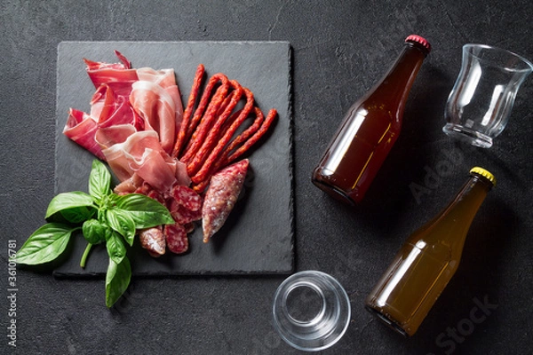 Fototapeta Prosciutto, sausage Fuet and chorizo, bottled beer on a black slate board. Antipasti concept.