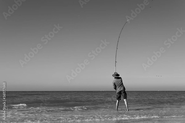 Fototapeta Black and white photo of a fisherman wearing a hat throwing his long curved fishing rod into the north sea in shorts standing barefoot in the water on Terschelling island , the Netherlands 