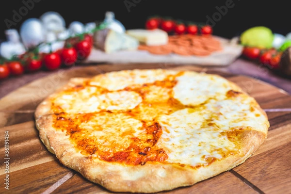 Fototapeta Homemade freshly baked Pizza Margherita with melted mozzarella on the top on a wooden board with lots of decorative vegetables. Concept of fresh hot pizza with free space for your text/decorations.