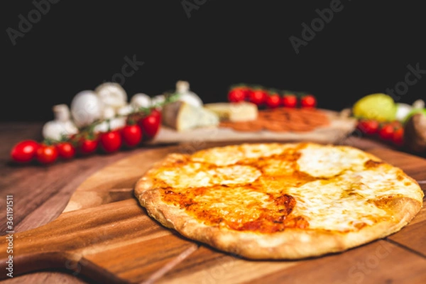 Fototapeta Homemade freshly baked Pizza Margherita with melted mozzarella on the top on a wooden board with lots of decorative vegetables. Concept of fresh hot pizza with free space for your text/decorations.