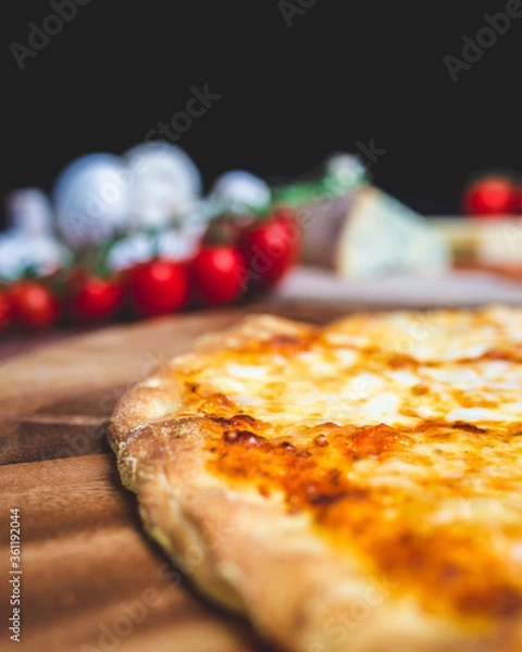Fototapeta Homemade freshly baked Pizza Margherita with melted mozzarella on the top on a wooden board with lots of decorative vegetables. Concept of fresh hot pizza with free space for your text/decorations.