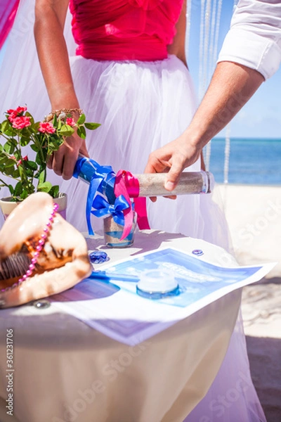 Fototapeta Bride and groom pouring colorful different colored sands into crystal vase close up during symbolic nautical decor destination wedding marriage ceremony on sandy beach in front of the ocean.Newlyweds 