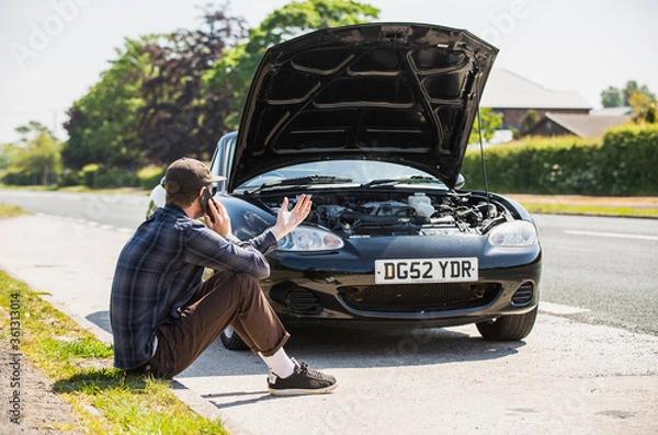 Obraz Young man next to his car, depressed and angry on the mobile phone telephone call after the car has broken down on the roadside waiting for assistance to arrive and recover or fix the vehicle