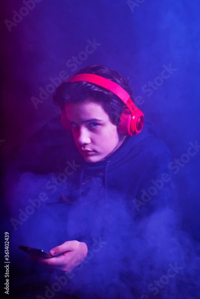 Fototapeta The guy in the headphones is playing on the phone. Thick smoke with red and blue tinges in the background. Dramatic contrasting light on the face as an artistic effect.