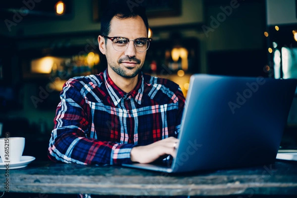 Obraz Portrait of smiling successful young man in optical eyeglasses updating software on digital laptop device connected to wireless 4G internet while looking at camera sitting in stylish coffee shop