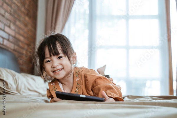 Fototapeta Asian little girl laughs happily when looking at the camera and holds a phone to watch videos
