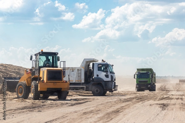 Fototapeta An excavator and dump trucks at the construction site outside the city. Road works on an intercity highway on a cloudy day