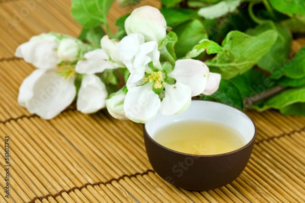 Fototapeta Green tea in a ceramic cup with branches of blossoming cherry tree on a bamboo background. Spring background.