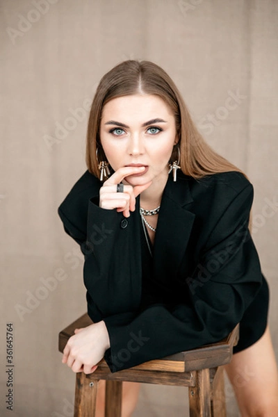 Fototapeta A beautiful girl of European appearance with brown hair, neutral makeup, in a black jacket looks into the frame with one hand near her face, a finger on her lips. Beige background.Vertical orientation