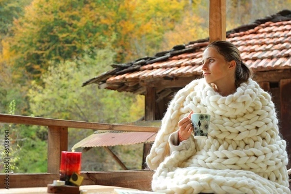 Fototapeta A young woman wrapped in a woollen blanket. She is drinking from a cup. It's a rural scene in a village.