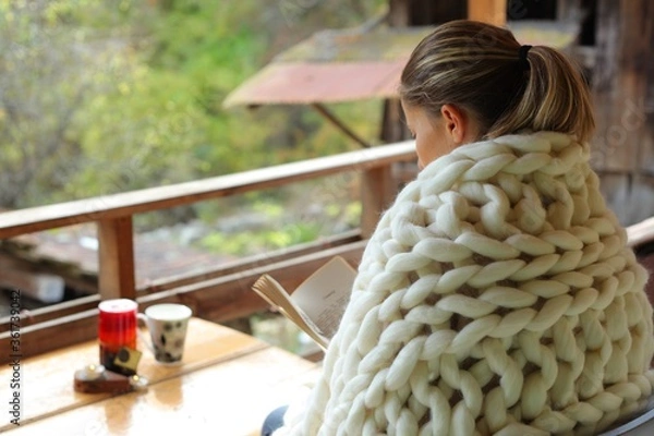 Fototapeta A young woman reading a book. She is cover in a woollen blanket. The photo is taken from behind her shoulder. It's a rural scene in a village.