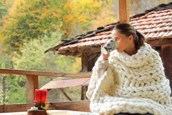 Fototapeta A young woman wrapped in a woollen blanket. She is drinking from a cup. It's a rural scene in a village.