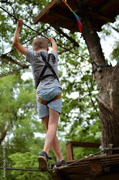 Fototapeta 
A child in equipment for climbers. A boy in an amusement park walks a tightrope with obstacles.