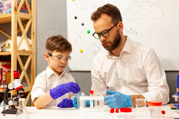 Fototapeta dad and son the child stayed at home conduct experiments on a white table, behind them on the wall is a white Board magnetic-marker