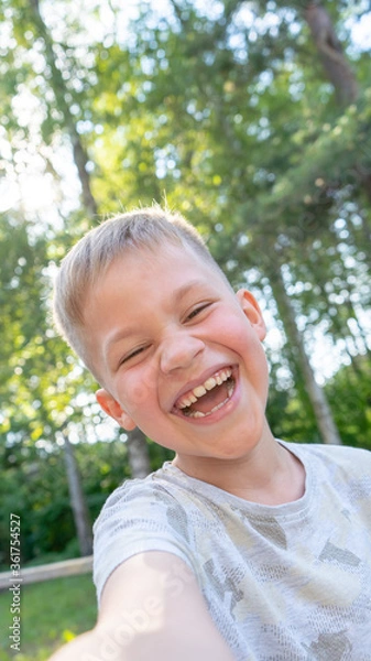 Fototapeta Authentic Funny small boy taking selfies and laughing. Smiling cute boy taking photos and facetime chatting with family. Vertical photo.