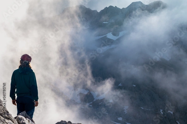 Fototapeta Montañero disfrutando de las vistas en la cumbre de una montaña de los picos de europa en un paisaje de nubes con las montañas de fondo y algo de nieve