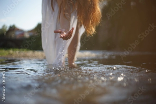 Fototapeta A girl/ young woman with red hair and a white dress. She is playing with water. The photo is taken in a rural scene. The weather is sunny, yet the light is soft and warm.