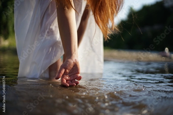 Fototapeta A girl/ young woman with red hair and a white dress. She is playing with water. The photo is taken in a rural scene. The weather is sunny, yet the light is soft and warm.