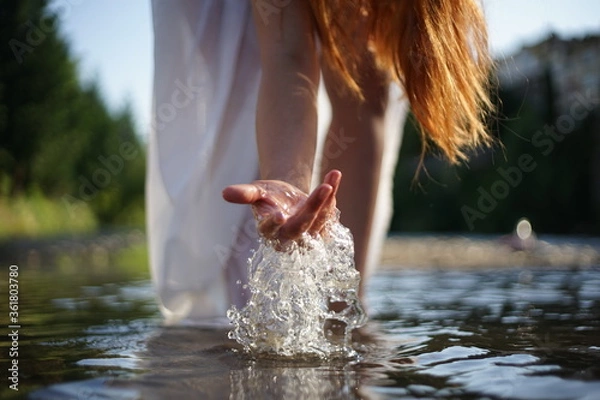 Fototapeta A girl/ young woman with red hair and a white dress. She is playing with water. The photo is taken in a rural scene. The weather is sunny, yet the light is soft and warm.