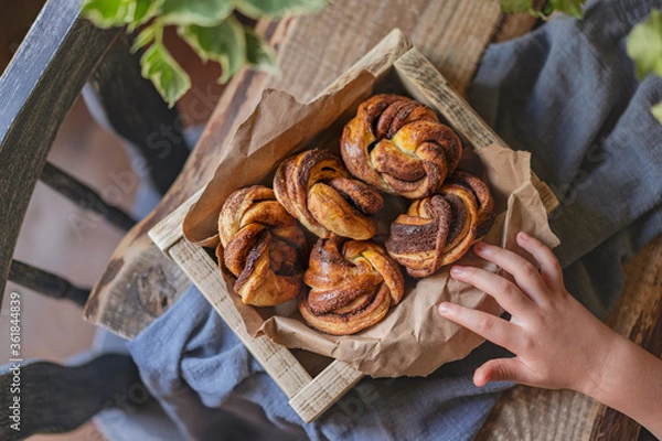 Fototapeta Cinnamon babka buns, homemade little babka pies in wooden box, rustic table with char, plant and child's small hand. Top view, flat lay, copy space. Kitchen with natural light, selective focus.