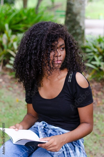 Obraz a young black woman between 20 and 30 years old sitting reading a book alone, in a park