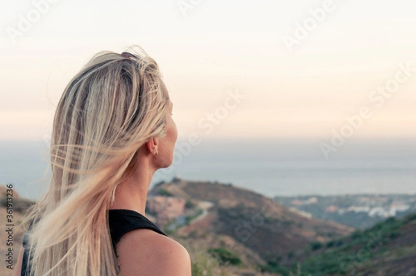 Fototapeta The view from the back of the head of a girl with hair fluttering  in the wind with a blurred background of hills and the Mediterranean Sea, looks dreamily into the distance.