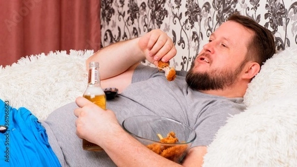 Fototapeta fat man drinks beer and eats unhealthy food chicken wings, bored in front of the TV outlook on the couch. The concept of malnutrition, quarantine at home, alcoholism.