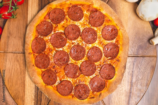 Fototapeta Pepperoni Pizza on a wooden pizza board with lots of ingredients and around. Concept of freshly homemade pizza with a lot of pepperoni salami on the top with free space for your text/decoration. 