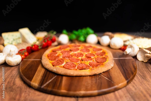 Fototapeta Pepperoni Pizza on a wooden pizza board with lots of ingredients and around. Concept of freshly homemade pizza with a lot of pepperoni salami on the top with free space for your text/decoration. 