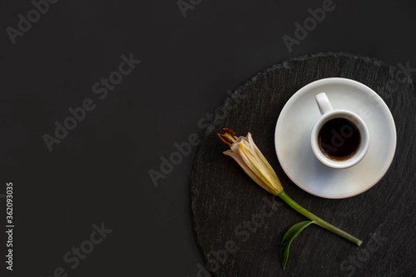 Obraz Cup of black coffee and fresh spring single bud of lily flower on a round slate on a black background. Romantic stylish elegance breakfast. Top view. Space for text.