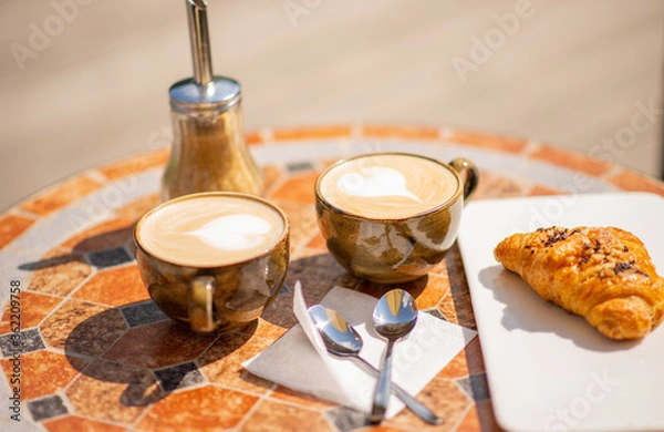 Fototapeta Two ceramic cappuccino cups and a croissant on a mosaic cafe table, on a Sunny summer day. Concept for meetings with friends, summer walks. Close-up, selective focus, horizontal position