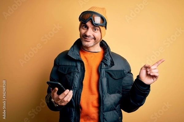 Fototapeta Young handsome skier man wearing ski goggles using smartphone over yellow background very happy pointing with hand and finger to the side