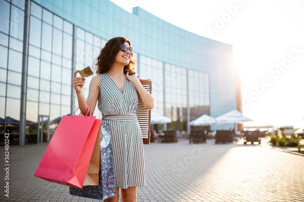 Obraz Young beautiful happy woman walks out of shopping mall with purchases. Shopaholic girl glad to buy clothes and stands outdoor with bags and credit card. Shop, sales, discount, spend money concept.