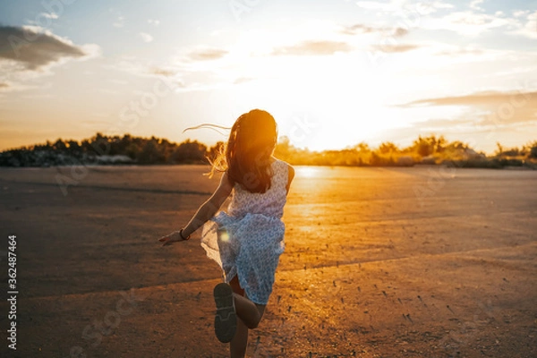 Fototapeta A little girl a child in a dress with long hair runs along an empty road in the sunset orange rays of the sun