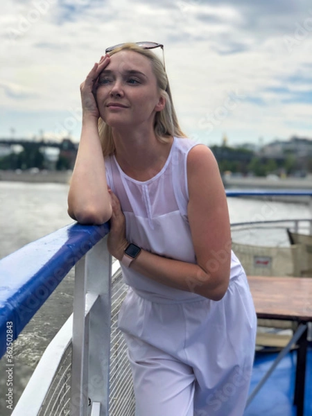 Fototapeta A beautiful woman stands aboard a small pleasure boat, leaning on the handrails. The blonde is resting on a pleasure boat. Smiling girl at the age of 28 years on the deck of the pleasure boat.