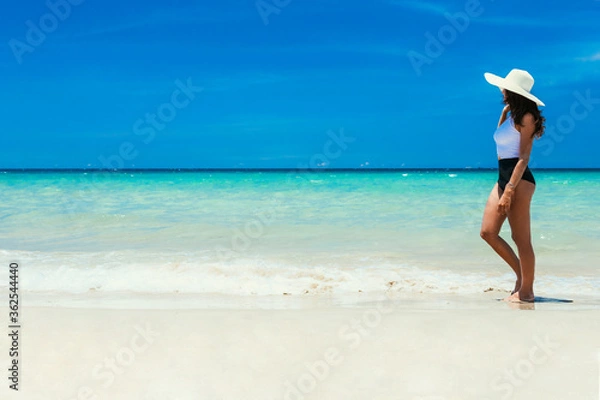 Obraz Sea, beach, a beautiful slender girl walks along the shore of a sandy beach with crystal clear bruise water in the sea in a straw hat