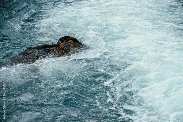 Fototapeta Mossy stone in azure water of mountain river close-up. Blue nature background with boulder with moss in turquoise water of mountain creek. Full frame of sea surf. Stone in ocean surf. Backdrop of tide