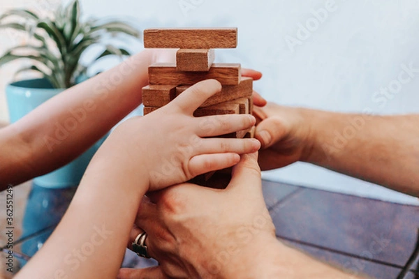 Obraz Father and child playing game tumble tower from wooden block. Man's and kids hands hold the tower together to prevent fall and crisis. Man helps child win. Concept of support and mutual assistance