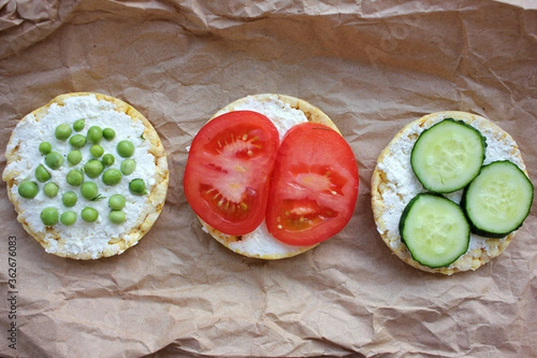 Fototapeta Round crispy corn sandwiches with vegetables toppings. Puffed corn cakes with cream cheese, tomatoes, cucumbers, and peas on craft paper texture. Healthy breakfast, snack, or diet concept. Top view