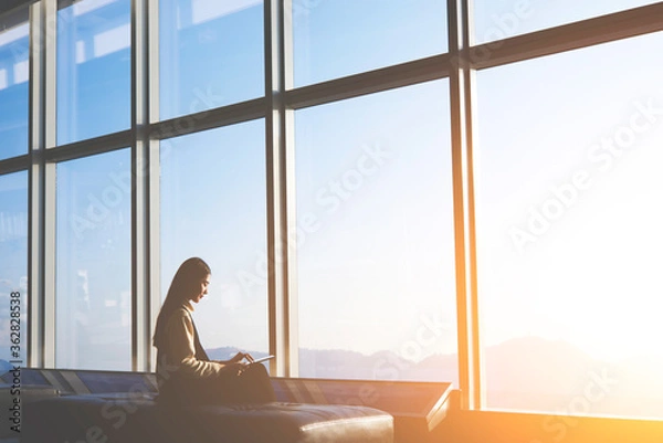 Fototapeta Silhouette of young asian woman is sitting in departure terminal of the airport building, while is using digital tablet for connect to wireless.Asian female entrepreneur is sitting front office window