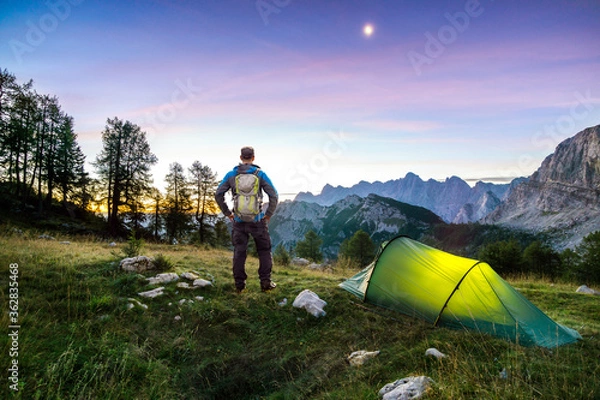 Fototapeta Hiker with Backpack standing on Mountain and Tent glowing under a moon night sky at sunset or sunrise twilight hour. Alps, Sleme, Triglav National Park, Slovenia.