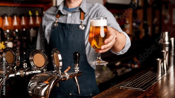 Fototapeta Fresh beer from keg. Glass of light lager with foam in hands of bartender in apron