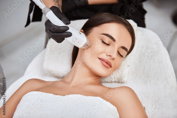 Fototapeta beautiful girl lying in spa salon covering breast with white towel and getting massage for her cheek with professional apparat