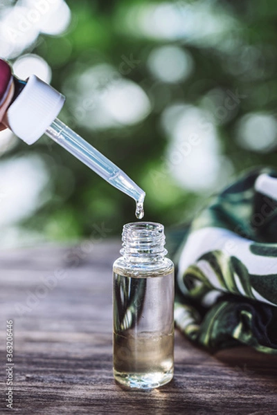 Fototapeta Essential oil falling from glass dropper. Dropper with aroma oil. Glass bottle of aroma oil isolated on wooden table. Spa Treatment.